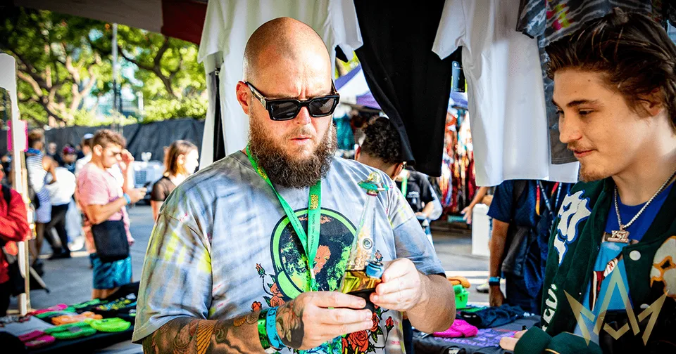 Two people at an outdoor market, one inspecting a colorful item while wearing sunglasses and a beard.