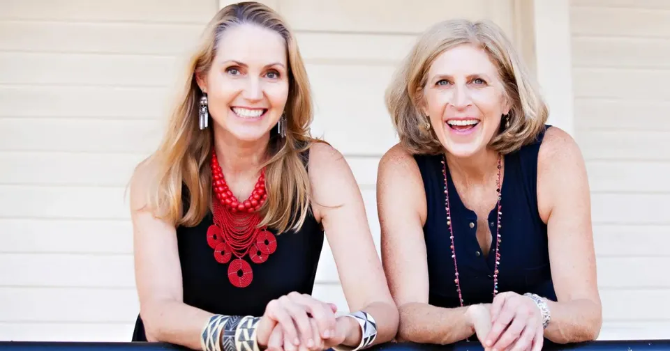 Two smiling women in black tops and colorful necklaces leaning on a railing against a light wall