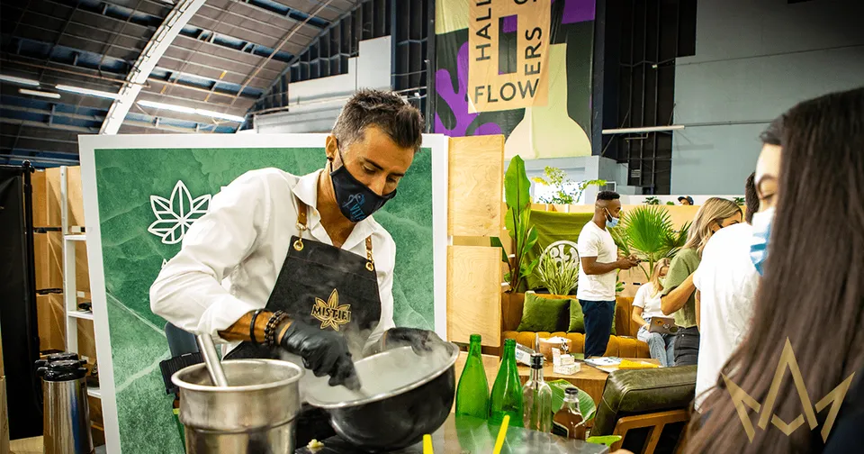 Chef demonstrating food preparation at a busy indoor market booth, stirring a pan with steam rising.