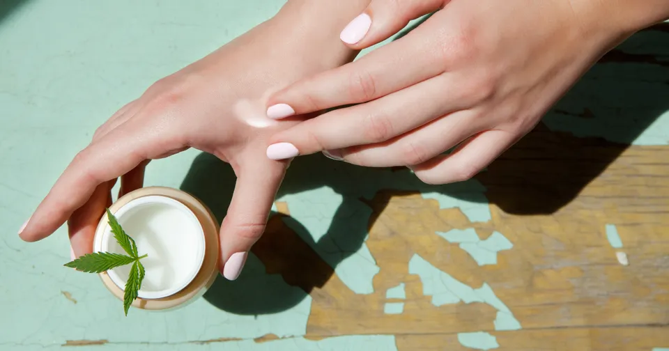 Hands applying cream beside a small jar on a sunlit table with leaf shadows