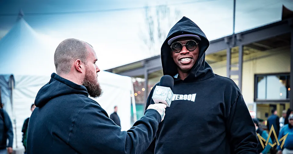 Man in black hoodie and sunglasses speaks into a microphone outdoors at an event, smiling.