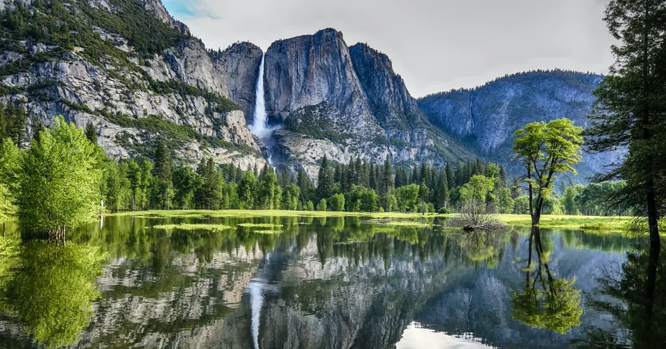 Mountain waterfall beside a calm lake, with green forest and rocky cliffs reflected in the water.