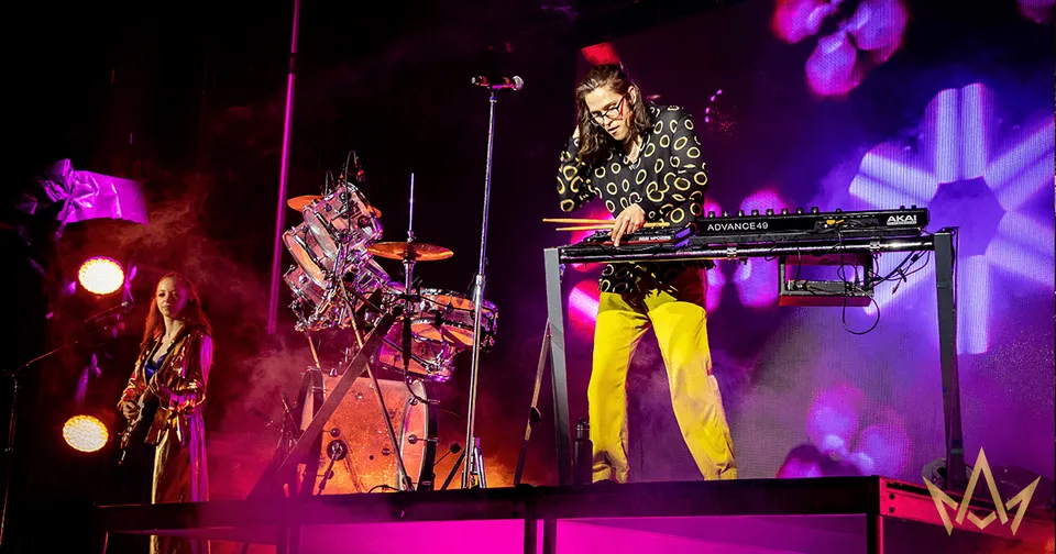 Musician playing keyboard onstage under pink and purple concert lights with drums in the background