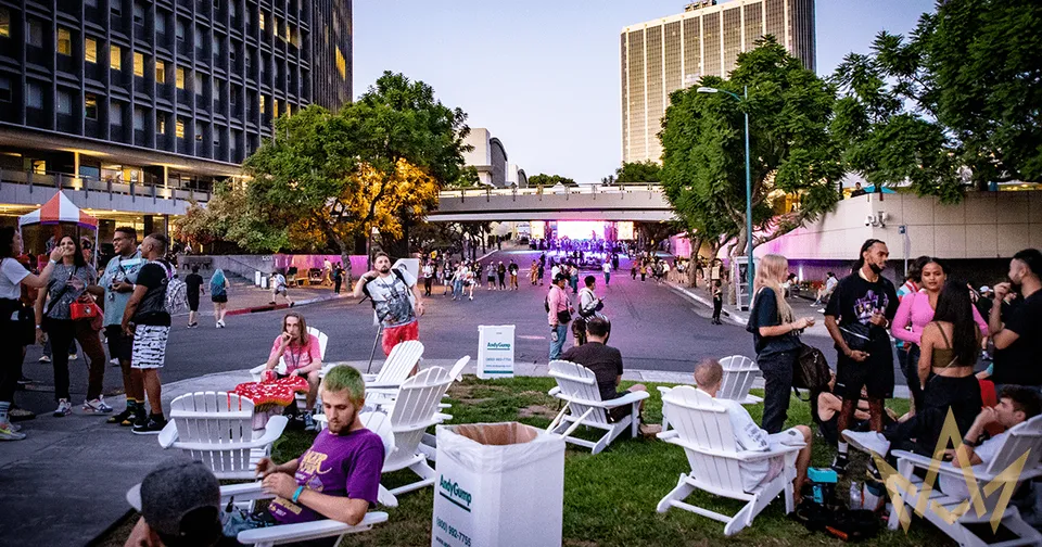 People relaxing and chatting in a city plaza at dusk, with white chairs and purple lighting in the background.