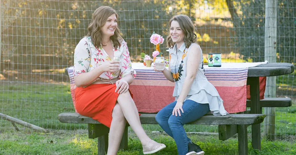 Two women sit at a picnic table outdoors with flowers and a red tablecloth, smiling and talking.