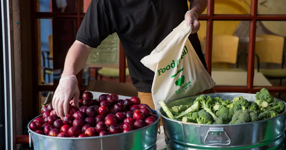 Person bagging groceries beside bins of red apples and green lettuce in a market