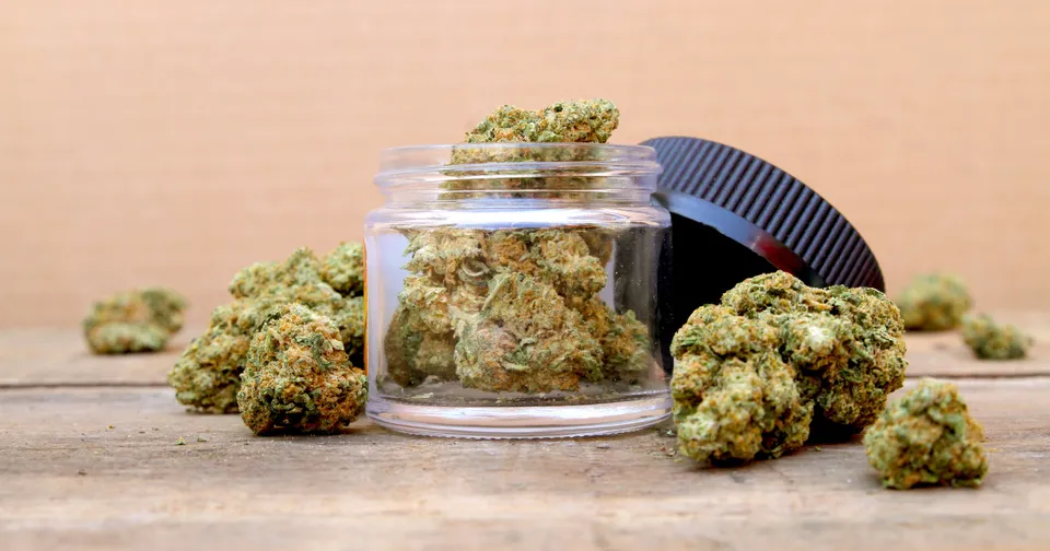 Cannabis buds in a glass jar on a wooden table, with more buds scattered nearby.