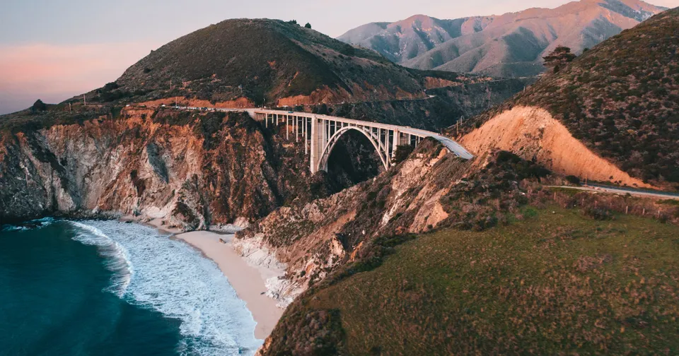Coastal bridge over a rocky cliff beside a sandy beach and blue ocean at sunset