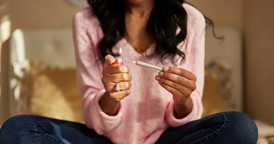 Person in a pink sweater holding a lighter and a syringe while sitting cross-legged on a couch