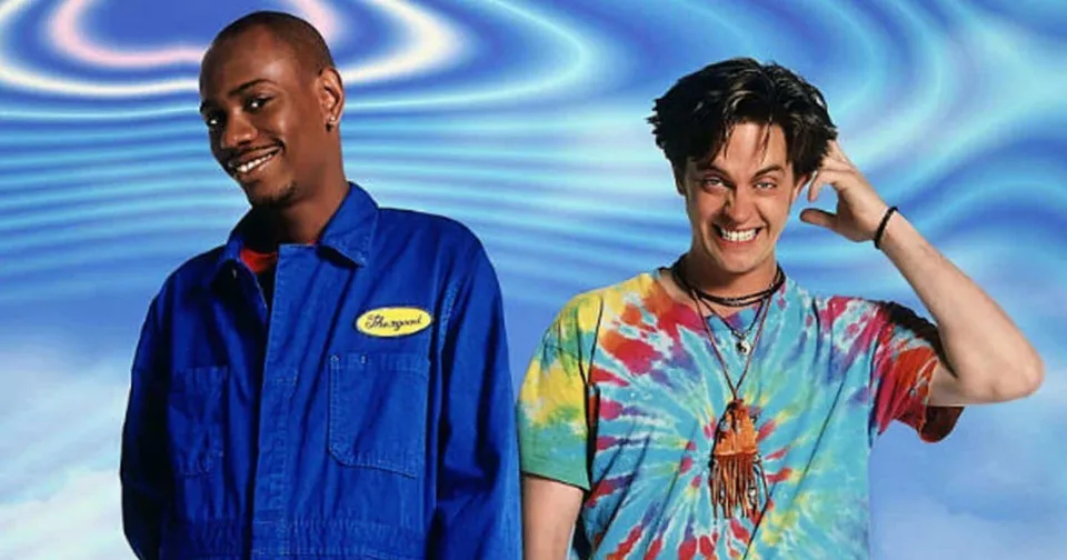 Two men posing against a blue wavy background, one smiling in a blue shirt and one in a tie-dye shirt scratching his head