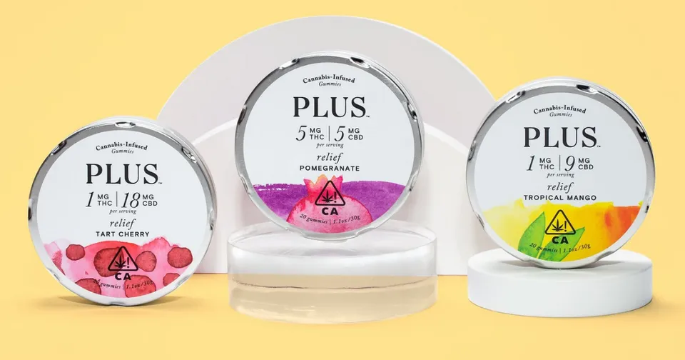 Three round PLUS compact cases in white, pink, and gray on clear stands against a yellow background