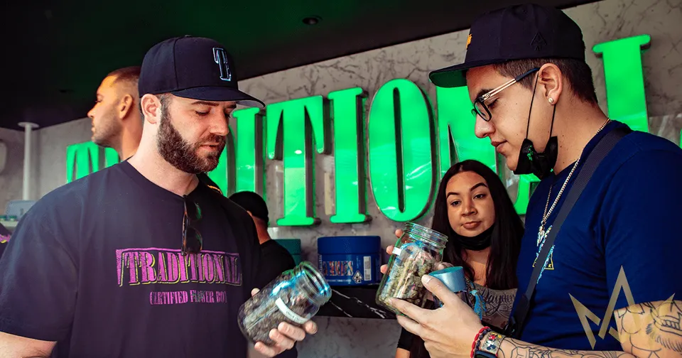 Two men and a woman at a table, examining jars in a neon-lit venue with large green letters behind them