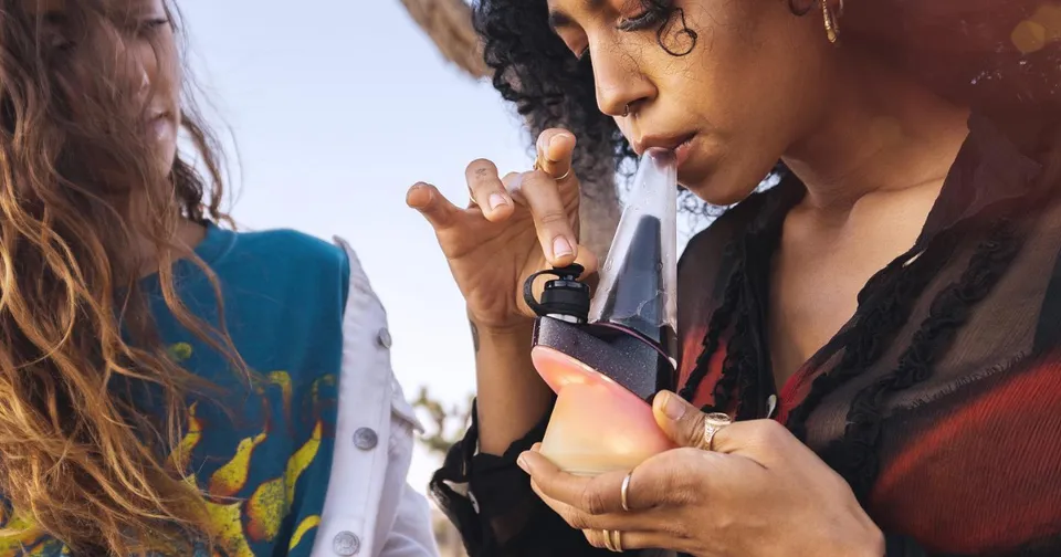Woman holds a small black device near her face while standing beside another person outdoors.