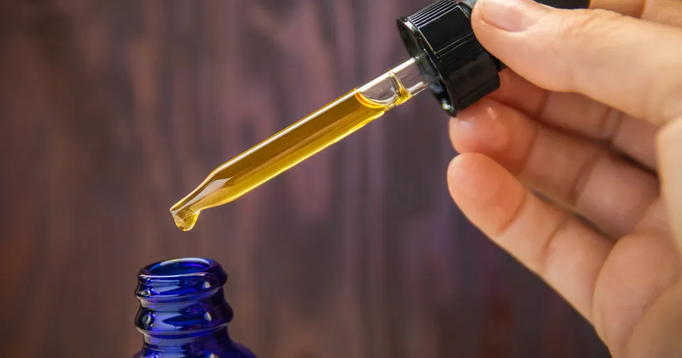 Hand holding a dropper with amber liquid above a blue glass bottle opening
