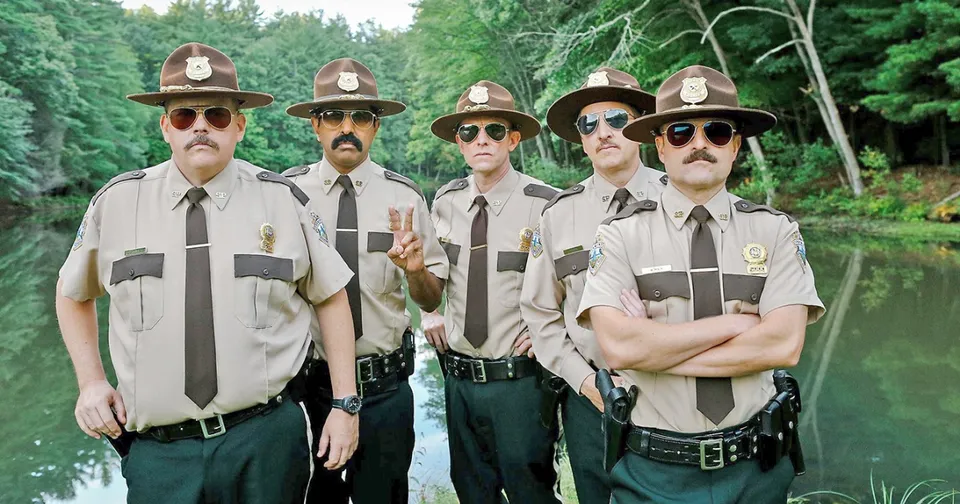 Five park rangers in tan uniforms and hats stand together outdoors in a green forest.