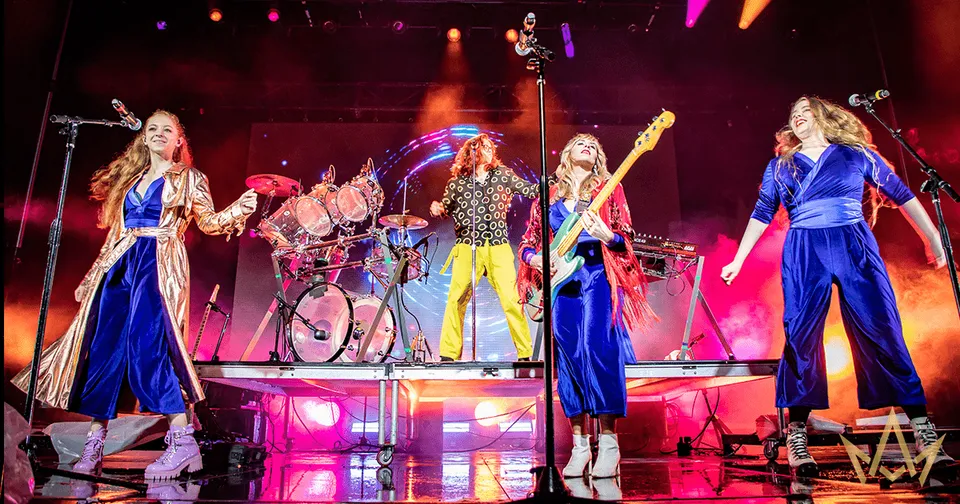 Four musicians perform on a brightly lit stage with red and purple lights, guitars, and drums.