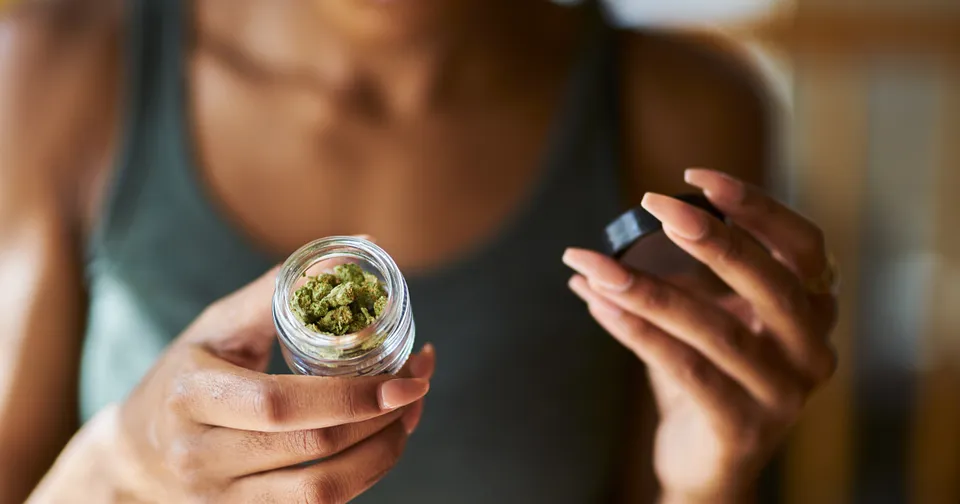 Person holding a rolled cannabis joint and a small black container indoors