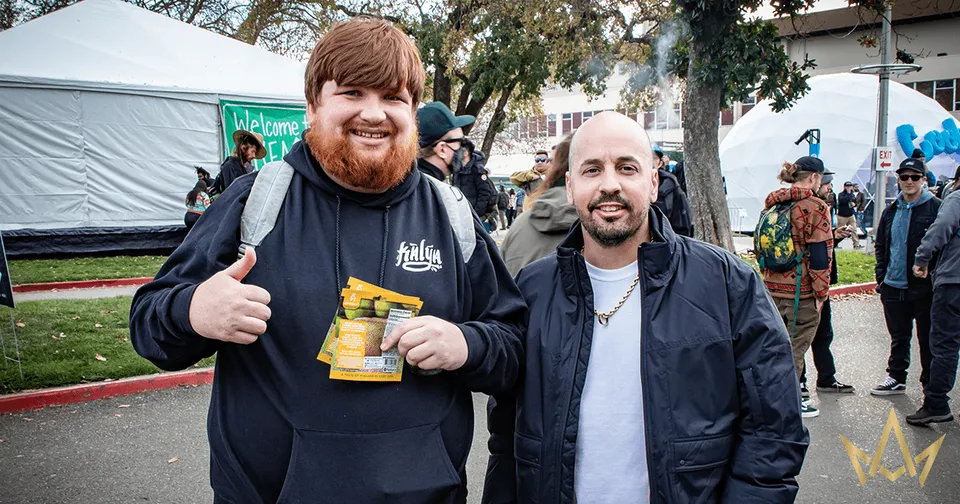 Two smiling people standing outdoors at an event, one holding a snack bag and giving a thumbs-up.