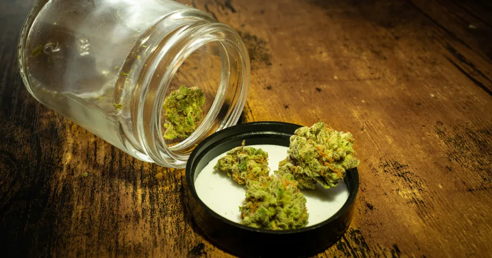 Glass jar spilling green cannabis buds onto a small black dish on a wooden table