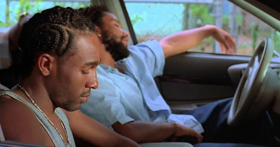 Two people sit in a car, one looking down in thought, the other leaning back with eyes closed.