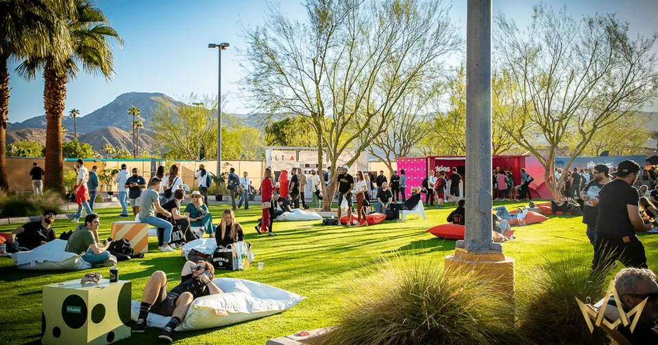 People relaxing on blankets at a sunny outdoor park gathering with trees, mountains, and pink food trucks.