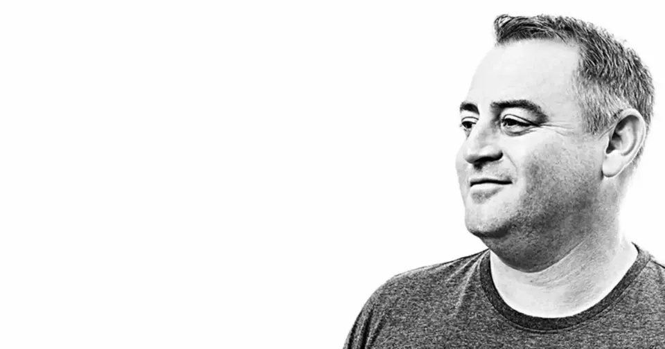 Black-and-white portrait of a smiling man in a dark T-shirt, facing right against a white background