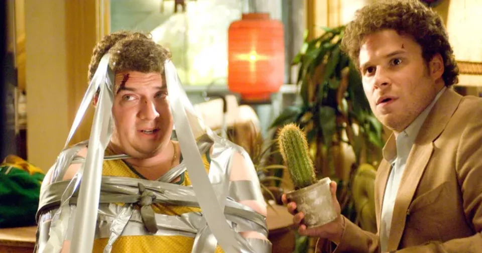 Two men in a kitchen, one wrapped in clear tape, the other holding a potted cactus with a surprised look.