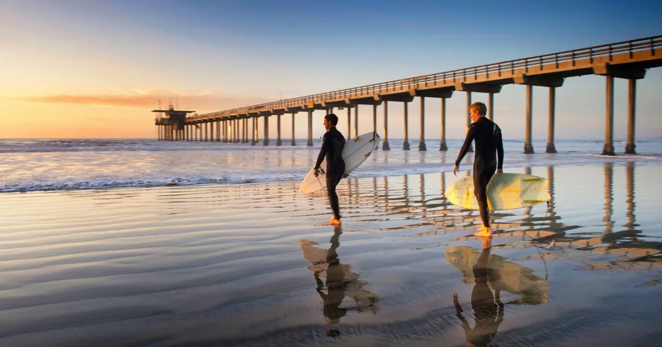 Two people wade in shallow water at sunset beside a long pier, with golden reflections on the wet sand.
