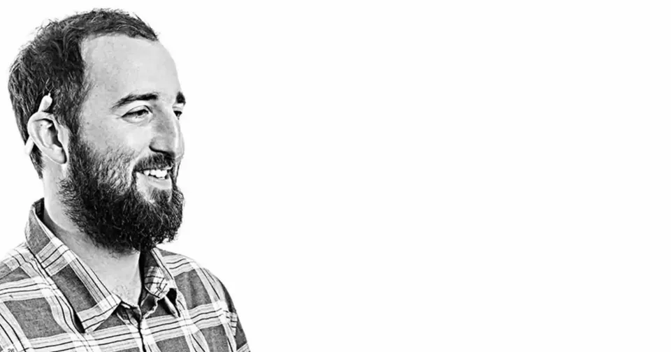 Black-and-white portrait of a smiling bearded man in a plaid shirt, facing left.