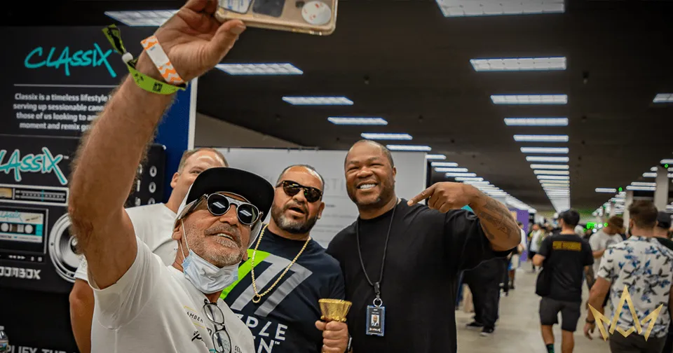 Three smiling people posing at an indoor event, one holding up a selfie stick in front of booths and lights.