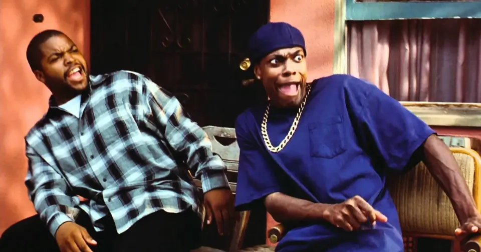Two people leaning back and laughing on a couch, one in a blue shirt and cap, one in a plaid shirt.