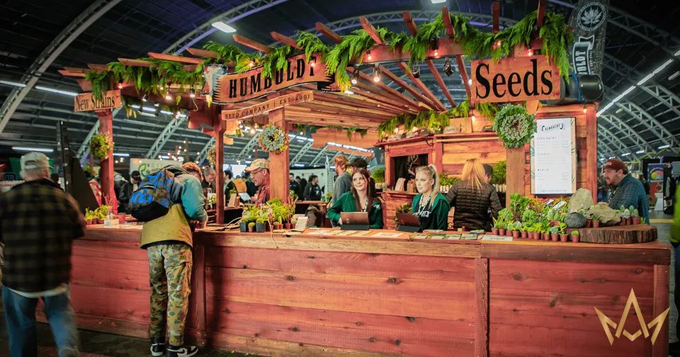 Rustic outdoor market stall labeled “Seeds,” with people browsing under warm string lights and greenery.