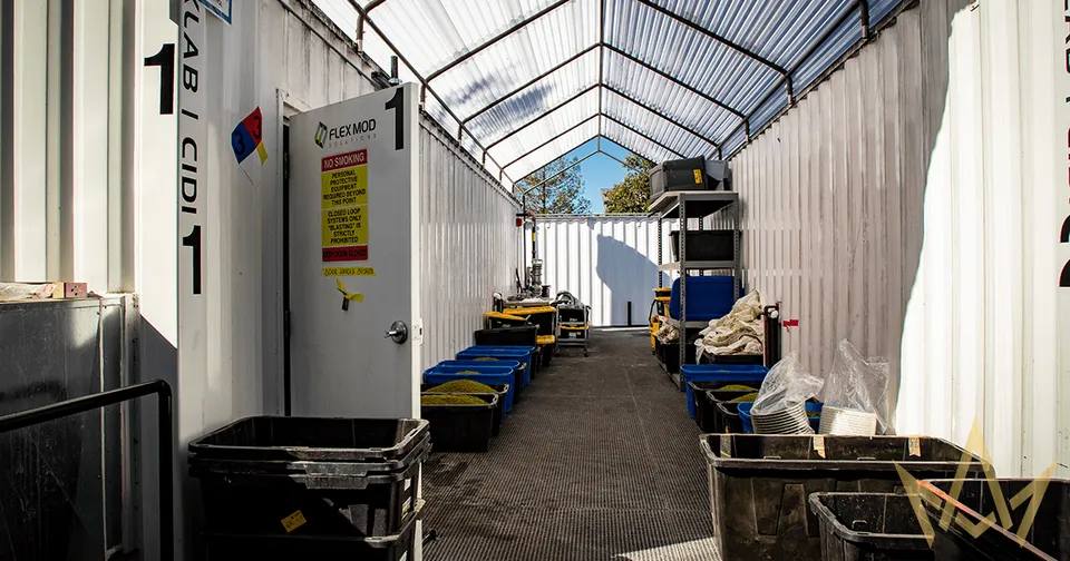 Narrow covered corridor with white walls, blue chairs, and yellow carts under a translucent roof