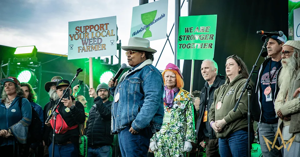Protesters holding signs at an outdoor rally on a wet day, wearing rain jackets and hats