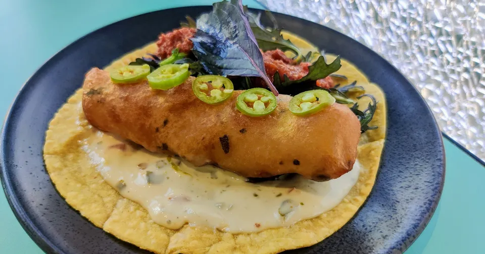 Breaded fish fillet on a blue plate with creamy sauce, sliced green peppers, and a yellow tortilla base.