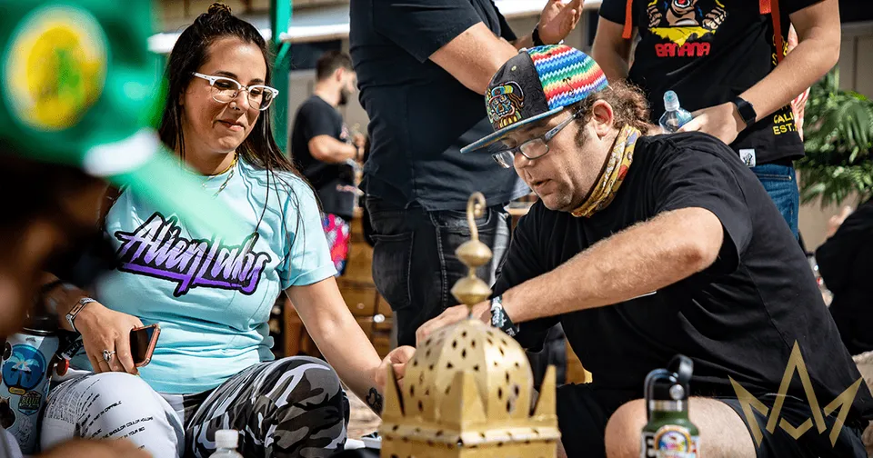 Two people seated outdoors, smiling while playing a wooden tabletop game with a gold crown-like piece.