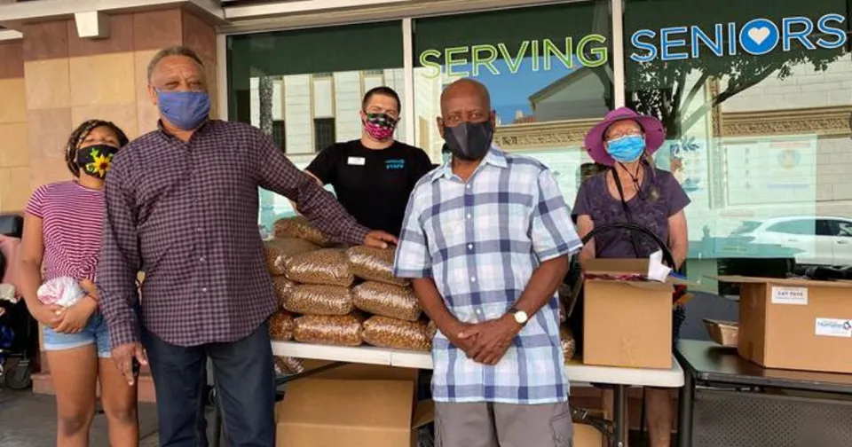 People wearing masks stand with stacks of bagged supplies outside a building labeled “SERVING SENIORS.”