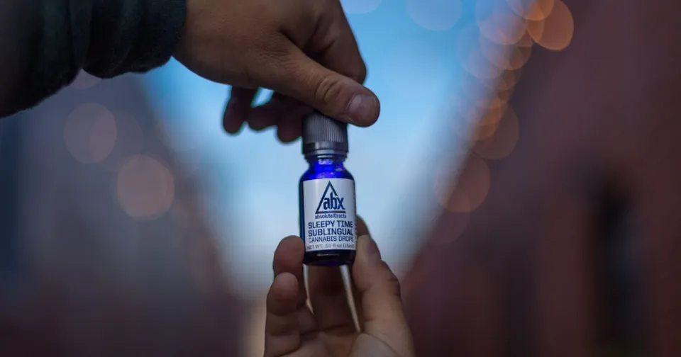 Hands holding a small blue vial with a label, against a blurred background of lights.