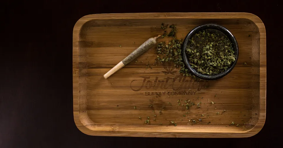 Wooden tray with a lit joint and a bowl of cannabis on a dark table