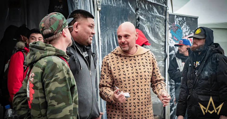 Several people in jackets stand outdoors, one man in a beige sweater holding an object while speaking.