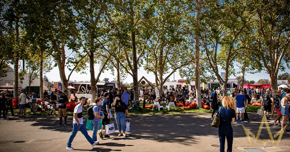 Crowd gathered under trees at an outdoor park event with people walking and sitting on benches