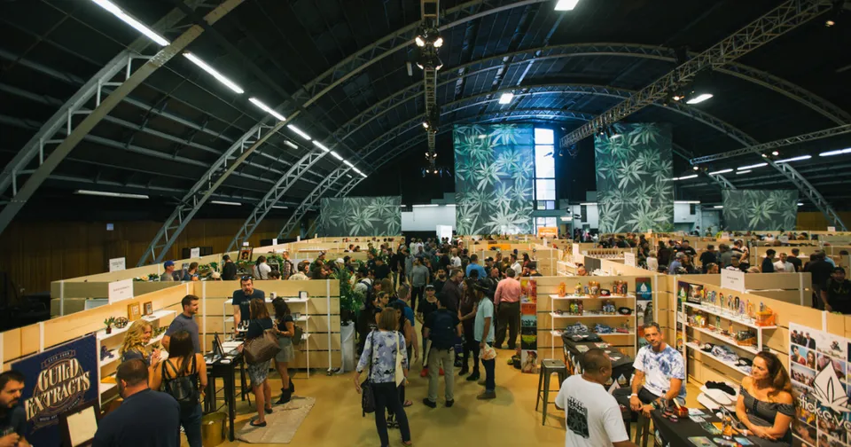 Indoor market or fair under a large arched roof, with people browsing wooden stalls and displays.